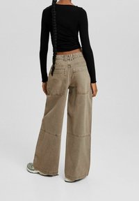 Wide-legged, beige cargo pants with prominent stitching and back pockets. Paired with a fitted black long-sleeve top.
