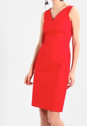 Donna con un vestito rosso senza maniche, lungo fino al ginocchio, con scollatura a V e orecchini pendenti, in piedi davanti a uno sfondo chiaro e semplice.