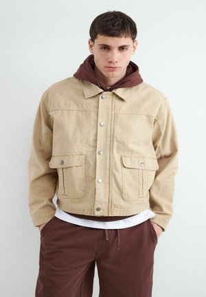 Młody mężczyzna w beżowej kurtce na guziki, pod którą ma brązową bluzę z kapturem oraz pasujące brązowe spodnie, stoi na jednolitym jasnym tle.