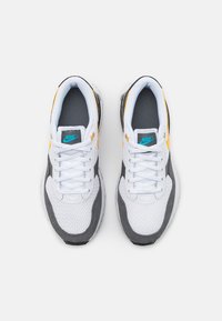 Sneakers Nike con una tomaia in rete bianca, accenti in suede grigio, dettagli gialli e un logo blu. Presentano un design con lacci e una suola testurizzata.
