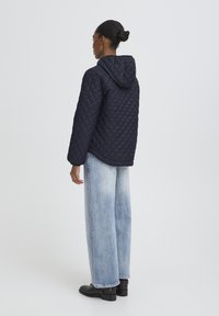 Mornarsko plava prošivena jakna s kapuljačom, s kutijastim oblikom. Kombinirana s svijetloplavim širokim trapericama i crnim gležnjačama.