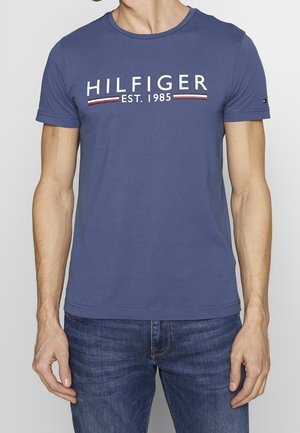 Mężczyzna w niebieskiej koszulce z krótkim rękawem z napisem "HILFIGER EST. 1985" na piersi oraz niebieskich dżinsach.