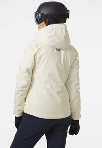 Crème ski-jack met verstelbare kap, slank ontwerp, waterdichte stof en een logo op de bovenrug. Zwarte handschoenen en donkere broeken completeren de look.