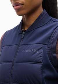 Κοντινό πλάνο ενός ατόμου που φοράει ένα σκούρο μπλε καπιτονέ γιλέκο Calvin Klein με μπροστινό φερμουάρ πάνω από καφέ μπλουζάκι.