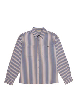 Jeune homme portant une chemise à boutons à rayures verticales bleu et beige, une casquette beige avec un logo foncé et un pantalon vert foncé, les mains dans les poches.