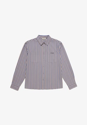 Junger Mann trägt ein blau-beiges, vertikal gestreiftes Hemd mit Knöpfen, beige Kappe mit dunklem Logo und dunkelgrüne Hose, Hände in den Taschen.