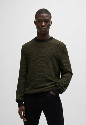 Maglione a maglia di colore verde scuro con righe orizzontali e un colletto e polsini a costine neri, indossato con pantaloni neri.