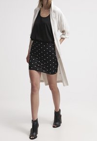 Débardeur noir associé à une jupe noire cintrée à pois blancs. Un long manteau léger beige clair complète la tenue. Bottines noires.