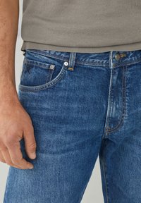 Vaqueros de mezclilla en un tono azul medio con un ajuste clásico. Las características incluyen una textura sutilmente desgastada, costuras en color naranja y cierre de botón metálico.