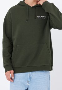 Felpa con cappuccio verde oliva realizzata in tessuto morbido, con tasca a marsupio, cappuccio con coulisse e scritta logo bianca sul petto a sinistra.