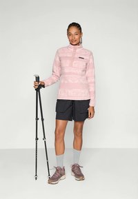 Rosa fleece-genser med geometriske mønstre, glidelås i kragen og svart logo. Kombinert med svarte cargo-shorts, grå sokker og brune turstøvler.