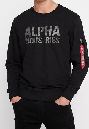 Mężczyzna w czarnej bluzie z kapturem z napisem kamuflażowym "ALPHA INDUSTRIES" na przodzie i czerwoną metką "REMOVE BEFORE FLIGHT" na kieszeni na lewym rękawie.