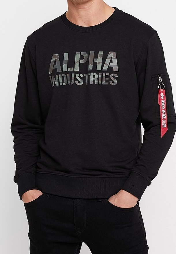 Homme portant un sweat-shirt noir avec le texte camo « ALPHA INDUSTRIES » à l'avant et une étiquette rouge « REMOVE BEFORE FLIGHT » sur la poche de la manche gauche.