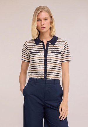 Donna bionda che indossa un top a maniche corte a righe in maglia con colletto blu navy e pantaloni blu navy a vita alta, in piedi con una mano in tasca.