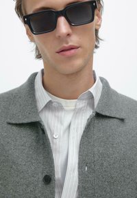 Giovane uomo che indossa occhiali da sole neri rettangolari, un cappotto di lana grigia e una camicia bianca con sottili righe verticali.