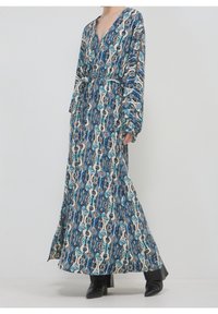 Robe longue avec un motif abstrait bleu, blanc et noir, manches longues, col en V, ceinture à la taille, portée avec des bottines noires.
