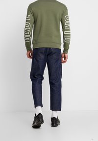 Sudadera verde oliva con texto blanco en las mangas, combinada con pantalones de mezclilla azul oscuro y zapatillas negras. Detalles visibles en los puños y la cintura.