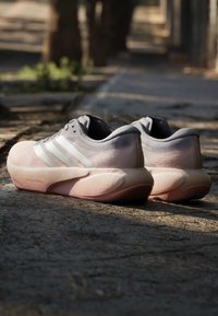 Ζευγάρι ανοιχτόροζ παπούτσια τρεξίματος Adidas με λευκές ρίγες, τοποθετημένα σε ηλιόλουστο υπαίθριο λιθόστρωτο.