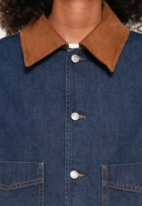 Spijkerjack met een bruine corduroy kraag, zilveren drukknoopjes, twee voorste zakken en gedetailleerde oranje stiksels op een donkerblauwe stof.
