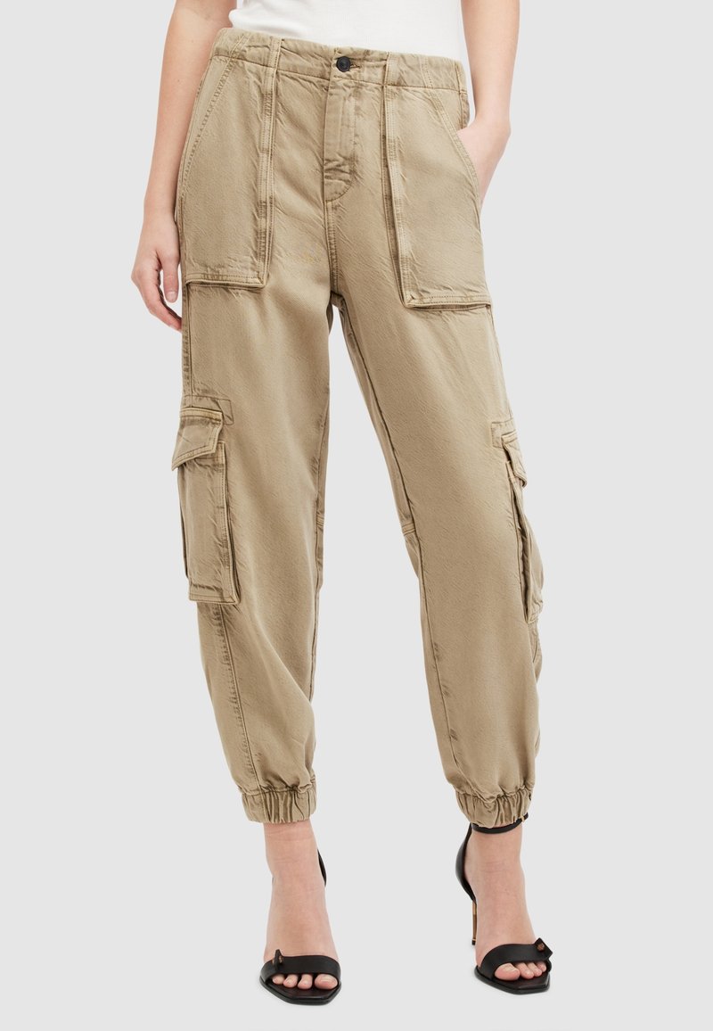 Pantalon cargo beige avec grandes poches sur le devant et les côtés, ourlets élastiqués aux chevilles, porté avec des talons ouverts noirs et un haut blanc.