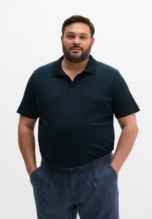 Mann mit kurzem dunklem Haar und Bart, der ein schwarzes kurzärmeliges Poloshirt und dunkle Hosen trägt, steht mit den Händen in den Taschen vor weißem Hintergrund.