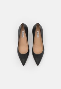 Escarpins à talons hauts en cuir noir avec un bout pointu, une texture lisse et une semelle intérieure nude arborant le logo "GUESS".