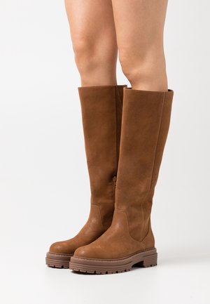 Bottes hautes en cuir marron avec semelles en caoutchouc épaisses portées sur des jambes nues, sur fond blanc uni.