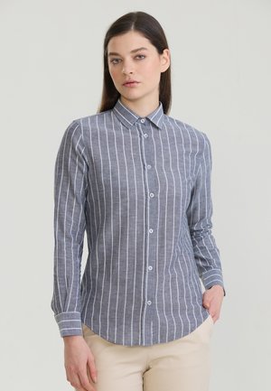 Młoda kobieta z długimi ciemnymi włosami, ubrana w szarą koszulę zapinaną na guziki z białymi pionowymi paskami i beżowe spodnie, stoi z jedną ręką w kieszeni.