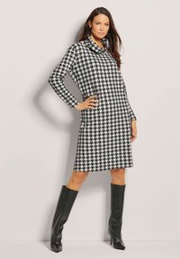 Schwarz-weißes Hahnentritt-Muster langärmeliges Kleid mit einem Cowl-Neck; kombiniert mit hohen schwarzen Lederstiefeln. Glatte Textur.