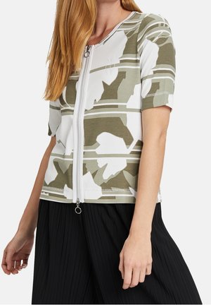 Femme portant un haut court zippé avec un motif abstrait vert et blanc, associé à une jupe noire plissée.