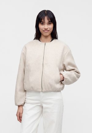 Junge Frau mit schwarzen Haaren, die eine beige Zip-up-Jacke und weiße Hose trägt, steht mit einer Hand in der Jackentasche vor einem schlichten Hintergrund.
