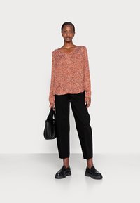Blusa de manga longa em V laranja com padrão floral em branco e preto; combinada com calças pretas de corte reto e mocassins pretos. Asegura uma bolsa preta.