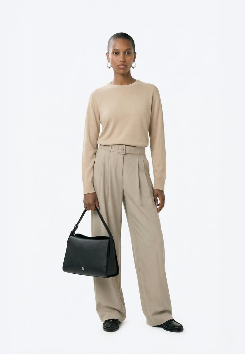 Donna in piedi, indossa un top beige a maniche lunghe e pantaloni a gamba larga a vita alta, tiene una borsa nera con una mano.