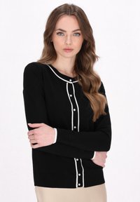Cardigan nero con bordi bianchi, presenta scollatura rotonda, maniche lunghe e bottoni decorativi. Materiale morbido con leggera elasticità, design aderente.