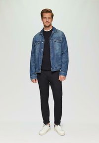 Denimjack in medium blauw, met twee borstzakken, gedragen over een zwarte trui. Gepaard met zwarte broeken en witte sneakers.