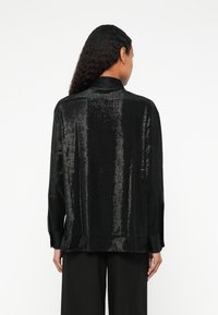 Maglietta nera a maniche lunghe con una texture lucida, caratterizzata da un colletto e una vestibilità rilassata, progettata con una parte posteriore dritta e spacco laterale.