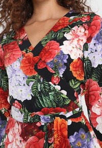 Robe portefeuille fleurie arborant des fleurs rouges, roses et violettes vibrantes sur un fond noir. Manches longues et ceinture assortie soulignent la taille.