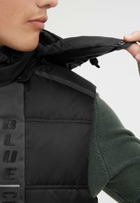 Schwarze gepolsterte Weste mit hohem Kragen, ausgestattet mit einem Reißverschluss und Druckknopfverschluss. Der strukturierte grüne Stoff, der am Arm sichtbar ist, sorgt für einen Kontrast.