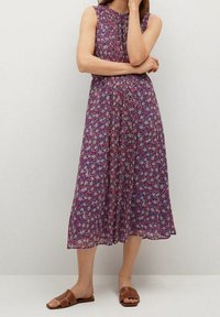 Robe sans manches violette avec un motif floral, longueur genoux, tissu léger, design plissé, portée avec des sandales plates marron.
