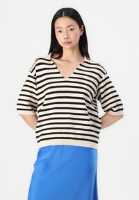 Pull en tricot rayé crème et noir avec un col en V et des manches courtes, associé à une jupe bleue fluide. Texture douce et coupe décontractée.