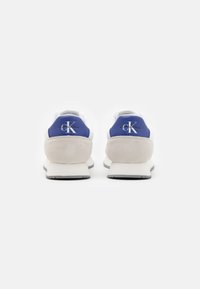 Zapatillas blancas y azules con una cápsula de talón de ante, parte superior de tela y suela de goma. El logotipo está grabado en la parte trasera en color plata.