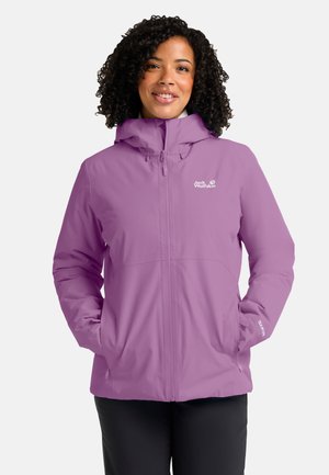 Frau mit lockigem Haar trägt eine violette Jack Wolfskin Jacke mit Kapuze und Händen in den Taschen, lächelt vor einem einfarbigen Hintergrund.