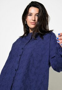 Camicia blu navy, con bottoni, realizzata in tessuto testurizzato, caratterizzata da ricami floreali e colletto alla mandarino, con maniche ampie.