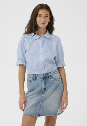 Junge Frau trägt eine hellblaue, geblümte Bluse mit Puffärmeln und einen ausgebleichten Jeansrock mit ausgefranstem Saum, steht vor weißem Hintergrund.