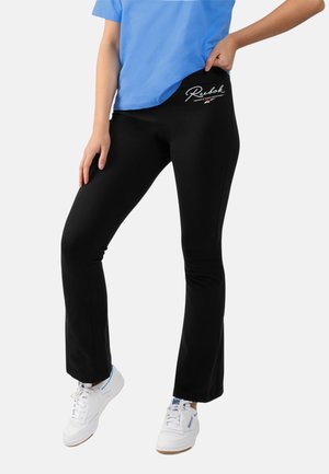 Persoon die zwarte Reebok legging, blauw shirt en witte Reebok sneakers draagt en met één hand op de heup staat.