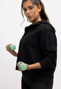 Felpa nera con zip anteriore, abbinata a pantaloni neri. Tieni in mano manubri verde chiaro. Tessuto morbido e design aderente.