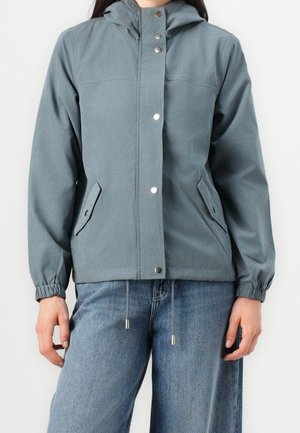 Persona con chaqueta azul grisácea con capucha, botones a presión, bolsillos frontales, puños elásticos y vaqueros azul.