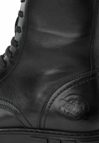 Bota de cuero negro con acabado suave, diseño de cordones y costuras visibles. Presenta un logo redondo en el lado y una suela resistente.