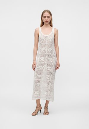 Jeune femme portant une robe midi blanche en crochet sans manches et des sandales beiges à talons hauts et à brides, debout devant un fond uni.