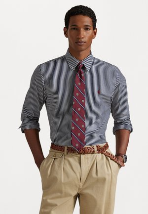 Chemise à rayures à manches longues en bleu marine et blanc, accompagnée d'une cravate bordeaux ornée de motifs de bouclier, de pantalons beiges et d'une ceinture tissée marron.